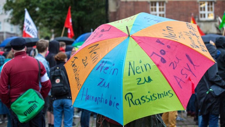 Foto eines bunten Regenschirms mit verschiedenen handschriftlichen Thesen. Jede Seite des Schirms hat einen Schriftzug. 3 davon sind auf dem Foto erkennbar: "Nein zu Rassismus", "Nein zu Homophobie", "Nein zu Faschismus"
