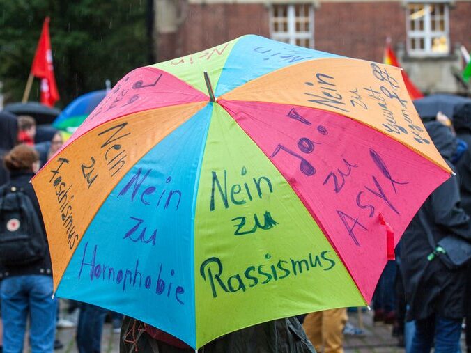 Foto eines bunten Regenschirms mit verschiedenen handschriftlichen Thesen. Jede Seite des Schirms hat einen Schriftzug. 3 davon sind auf dem Foto erkennbar: "Nein zu Rassismus", "Nein zu Homophobie", "Nein zu Faschismus"