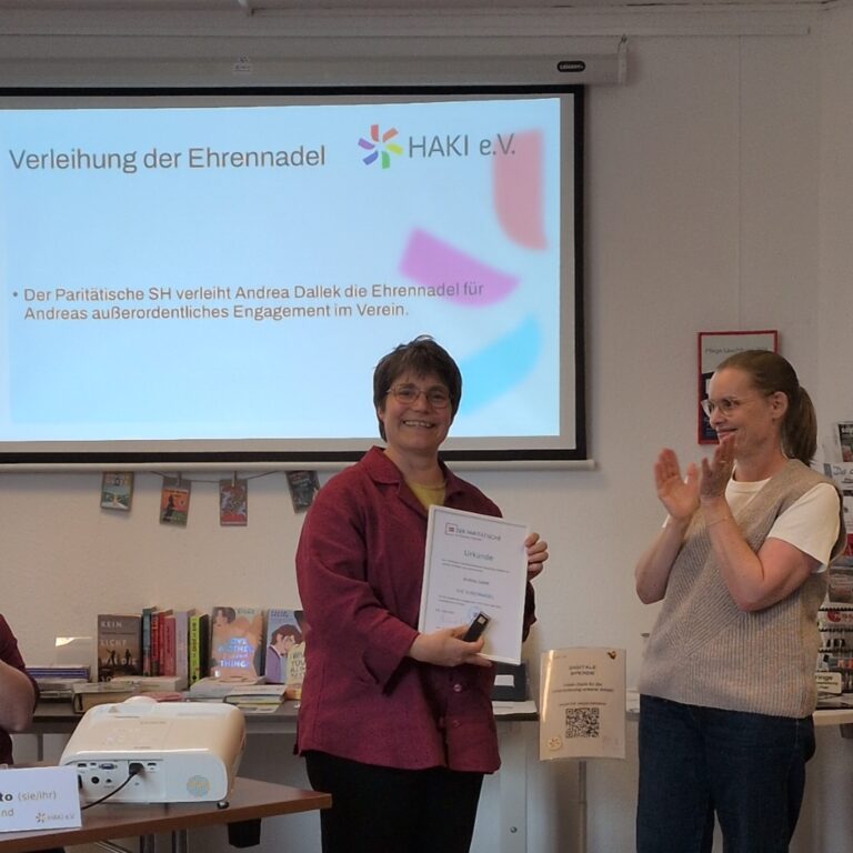 Links im Bild ist Andrea Dallek [keine Pronomen] zu sehen. Andrea hält eine Urkunde und die Ehrennadel und lächelt. Rechts daneben steht Prof. Dr. Melanie Groß für den Paritätischen SH und applaudiert.